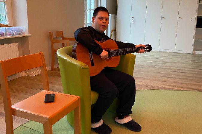 Junge mit Down-Syndrom sitzt in einem grünen Sessel und spielt Gitarre. Er trägt einen schwarzen Hoodie und Hausschuhe.