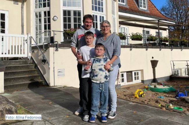 Familie steht lächelnd vor einem großen, alten Gebäude mit Fenstern und einem Gartenbereich. Zwei Kinder und zwei Erwachsene sind abgebildet.