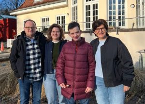 Vier Personen stehen zusammen, zwei Erwachsene und zwei Jugendliche, vor einem Gebäude mit großen Fenstern und einer Terrasse.