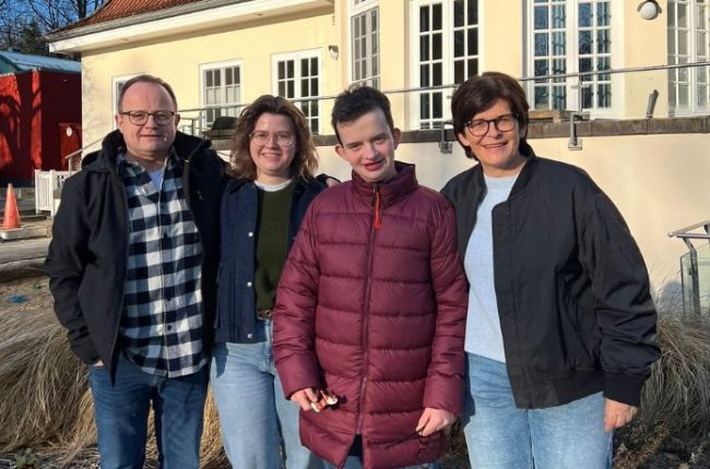 Vier Personen stehen zusammen, zwei Erwachsene und zwei Jugendliche, vor einem Gebäude mit großen Fenstern und einer Terrasse.
