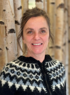 Frau mit lächelndem Gesicht und schwarzem, gemustertem Pullover vor einer hellen, strukturierten Wand aus Birkenstämmen.