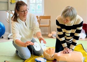Hände für Kinder: Erste-Hilfe-Kurs mit Ehrenamtlichen – Kursleiterin platziert Defibrillator-Pads an einer Übungs-Puppe, während eine Teilnehmerin aus dem Ehrenamt Herzdruckmassage übt.