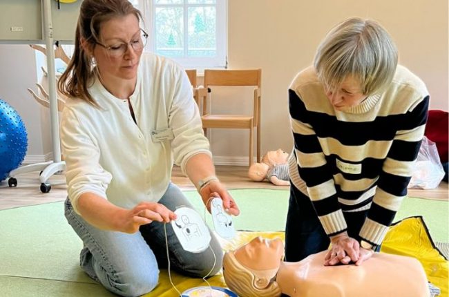 Hände für Kinder: Erste-Hilfe-Kurs mit Ehrenamtlichen – Kursleiterin platziert Defibrillator-Pads an einer Übungs-Puppe, während eine Teilnehmerin aus dem Ehrenamt Herzdruckmassage übt.