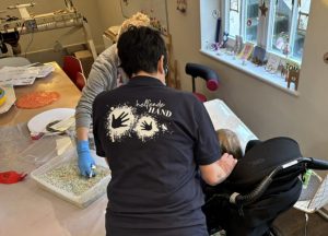 Claudia in einem dunkellblauen Shirt mit dem Logo 'Helfende Hand' und zwei Handabdrücken auf dem Rücken, während Mitarbeitende und eine Person im Rollstuhl in einer Werkstatt arbeiten; bunte Materialien liegen auf dem Tisch.