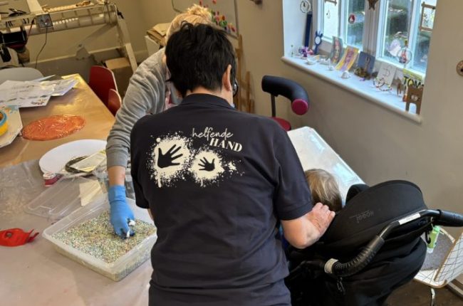 Claudia in einem dunkellblauen Shirt mit dem Logo 'Helfende Hand' und zwei Handabdrücken auf dem Rücken, während Mitarbeitende und eine Person im Rollstuhl in einer Werkstatt arbeiten; bunte Materialien liegen auf dem Tisch.