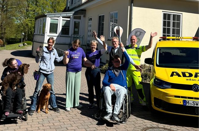 Beim Kinder-Lebens-Lauf 2026 winken Menschen vor gelbem ADAC-Auto mit Engelsfackel im Neuen Kupferhof.