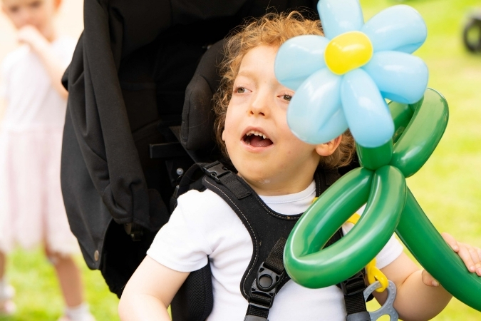 Ein kleiner Junge sitzt in einem Buggy und freut sich über seine blau-grüne Luftballonblume, di er in seiner Hand hält.