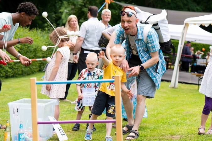 Zwei kleine Jungs spielen mit ihrem Vater Leitergolf und sind gerade dabei zu werfen. Im Hintergrund tummeln sich andere Menschen.