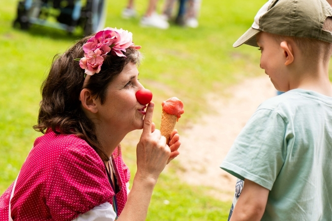 Eine Frau mit dunklen kurzen Haaren und pinkem Clownskostüm und Blumen in den Haaren ist von der Seite zu sehen. Sie hält ein Erdbeereis mit Waffel in der Hand und schaut einen kleinen blonden Jungen mit Basecap und hellgrünem Shirt an.
