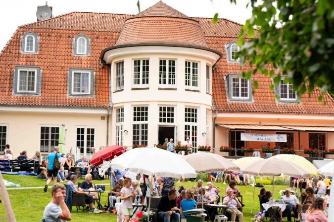 Menschen sitzen im Garten des Neuen Kupferhofes unter Sonnenschirmen. Im Hintergrund ist das Haupthaus zu sehen.