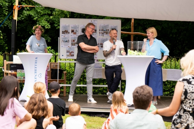 Zwei Männer stehen mittig auf der Bühne. Einer von ihren hält ein Mikrofon und einen Loszettel in der Hand. Zwei Frauen stehen links und rechts neben ihnen.