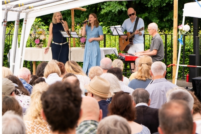 Zwei Frauen stehen vor Publikum und singen. Rechts von ihnen spielt ein Mann am Klavier und ein anderer Mann die Gitarre.