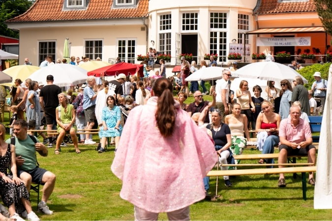 Eine Sängerin im rosa ABBA-Kostüm singt vor Publikum. Sie ist von Hinten zu sehen, die Menschen gucken sie an.