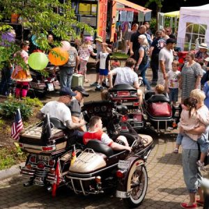Beim inklusiven Midsommarfest 2026 sitzen Gäste in Sidecar-Motorrädern, während Kinder mit Ballons und Familien den festlichen Garten füllen.