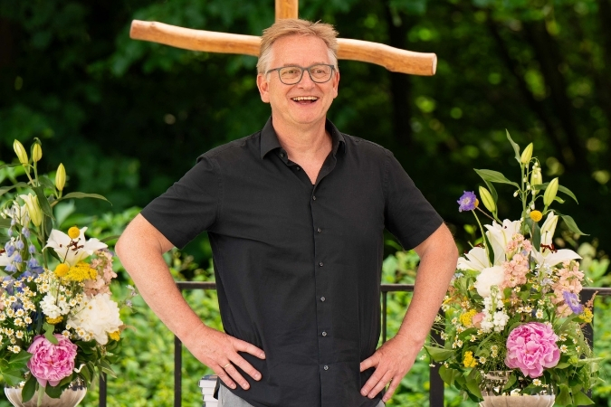 Steffen Schumann steht im schwarzen Hemd auf der Bühne und lächelt. Links und Rechts von ihm sind Blumensträuße, im Hintergrund ein modernes Holzkreuz.