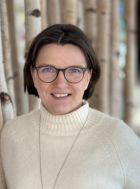 Mit Brille und hellem Rollkragenpullover steht Iris Drost vor einem unscharfen Holzuntergrund und blickt freundlich in die Kamera.