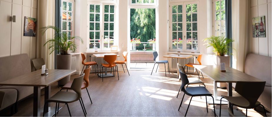 Im hellen Speisesaal  stehen Tische und Stühle neben großen Fenstern, dazu Pflanzen und warmes Tageslicht.