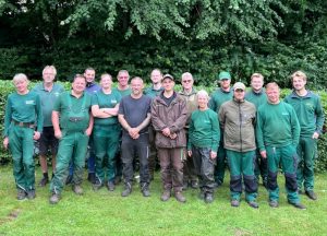 16 ehrenamtliche Friedhofsgärtner:innen stehen draußen auf einer Wiese vor der Hecke, die den Neuen Kupferhof umgibt.