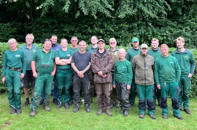 16 ehrenamtliche Friedhofsgärtner:innen stehen draußen auf einer Wiese vor der Hecke, die den Neuen Kupferhof umgibt.