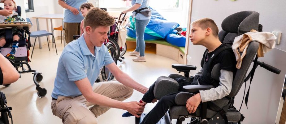 Ein männliche Pflegekraft hilft einem Jungen im Rollstuhl beim Socken anziehen; weitere Kinder und Pflegekräfte sind im Hintergrund zu sehen.