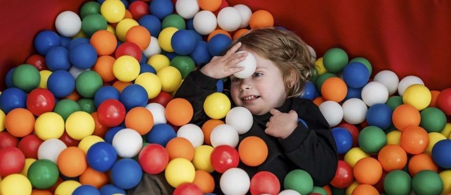 Ein lächelndes Mädchen liegt in einem bunten Bällebad und hält einen weißen Ball an die Stirn.